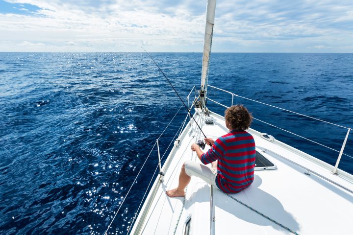 Person fishing on a sailboat in open sea under a cloudy sky.
